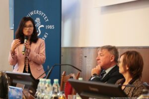 Po lewej stojąca prelegentka III Międzynarodowego Kongresu Języka i Kultury Polskiej trzymająca mikrofon. Za nią granatowy rollup z białym okrągłym logiem Uniwersytetu Szczecińskiego. Po prawej siedzący prorektor ds. nauki US i druga prelegentka.