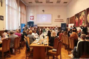 Na pierwszym planie siedzący przy stołach tyłem i bokiem do kadru uczestnicy III Międzynarodowego Kongresu Języka i Kultury Polskiej. Obserwują występ kilkuosobowego Zespołu Pieśni i Tańca Szczecinianie. W tle po lewej rollupy, mównica, nad nią godło Polski, dalej stół prezydialny. U góry na ścianie ekran, obok niego czerwony napis In unitate potentia, in potentia posteritas, po prawej portrety rektorów w gronostajach.