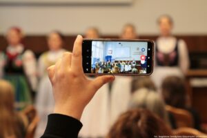 Na pierwszym planie ekran telefonu, który nagrywa występ kilkuosobowego Zespołu Pieśni i Tańca Szczecinianie, ubranego w ludowe stroje.