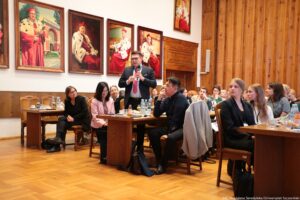 Ujęcie bokiem. Siedzący przy stołach (3 rzędy) uczestnicy III Międzynarodowego Kongresu Języka i Kultury Polskiej. W środku stojący mężczyzna z mikrofonem zadający pytanie podczas dyskusji. W tle na ścianie portrety rektorów w czerwonych togach z gronostajami.