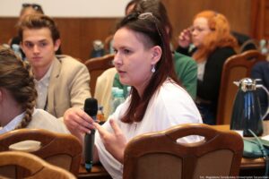 Ujęcie bokiem. Siedzący przy stołach (3 rzędy) uczestnicy III Międzynarodowego Kongresu Języka i Kultury Polskiej. Na pierwszym planie kobieta z mikrofonem zadająca pytanie podczas dyskusji.