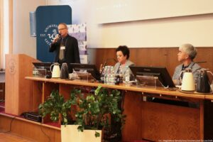 Po lewej stojący prelegent III Międzynarodowego Kongresu Języka i Kultury Polskiej trzymający mikrofon. Za nim granatowy rollup z białym okrągłym logiem Uniwersytetu Szczecińskiego. Po prawej prowadząca i drugi prelegent.