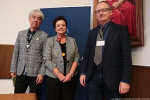 Dwóch prelegentów III Międzynarodowego Kongresu Języka i Kultury Polskiej i prowadząca tę część wydarzenia (w środku). Za nimi granatowy rollup z białym okrągłym logiem Uniwersytetu Szczecińskiego.