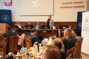 Na pierwszym planie siedzący przy stołach tyłem do kadru uczestnicy III Międzynarodowego Kongresu Języka i Kultury Polskiej. W tle po lewej granatowy rollup z białym okrągłym logiem Uniwersytetu Szczecińskiego, mównica, dalej stół prezydialny, za którym siedzą rektorzy. Pierwsza z prawej – prorektor ds. organizacji – stoi z mikrofonem i wita gości. W prawym górnym rogu na ścianie czerwony napis In unitate potentia, in potentia posteritas.