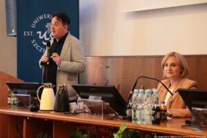 Po lewej stojący prelegent III Międzynarodowego Kongresu Języka i Kultury Polskiej trzymający mikrofon. Za nim granatowy rollup z białym okrągłym logiem Uniwersytetu Szczecińskiego. Po prawej prowadząca.