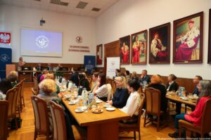 Ujęcie bokiem. Na pierwszym planie siedzący przy stołach tyłem i bokiem do kadru uczestnicy III Międzynarodowego Kongresu Języka i Kultury Polskiej. W tle po lewej granatowy rollup z białym okrągłym logiem Uniwersytetu Szczecińskiego, mównica, nad nimi godło Polski, dalej stół prezydialny, za którym siedzą rektorzy. Na ścianie ekran, a obok czerwony napis In unitate potentia, in potentia posteritas. Po prawej portrety rektorów w gronostajach.