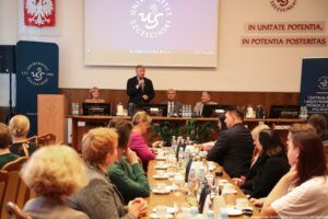 Na pierwszym planie siedzący przy stołach tyłem do kadru uczestnicy III Międzynarodowego Kongresu Języka i Kultury Polskiej. W tle po lewej granatowy rollup z białym okrągłym logiem Uniwersytetu Szczecińskiego, mównica, nad nimi godło Polski, dalej stół prezydialny, za którym siedzą rektorzy. Prorektor ds. nauki – stoi z mikrofonem i wita gości. U góry ekran, a obok na ścianie czerwony napis In unitate potentia, in potentia posteritas.