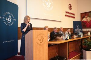 Po lewej granatowy rollup z białym okrągłym logiem Uniwersytetu Szczecińskiego. Dalej środku drewniana mównica, za którą stoi z mikrofonem dr Ewa Hendryk podczas III Międzynarodowego Kongresu Języka i Kultury Polskiej. Dalej stół prezydialny z rektorami US. U góry ekran, obok na ścianie czerwony napis In unitate potentia, in potentia posteritas, dalej portret rektora w gronostajach.