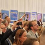 Zbliżenie na widownię nagrywającą telefonami wystąpienie na debacie Forum Ekonomicznego. W tle – na ścianie – plakaty promujące konkurs Matematyka w obiektywie i zdjęcia stanowiące wystawę.