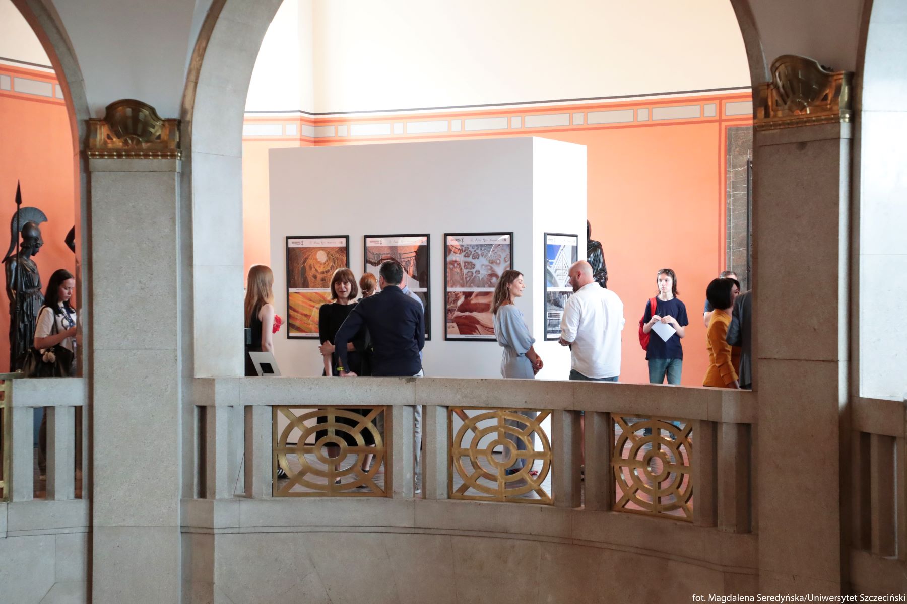 Wnętrze eleganckiej galerii lub muzeum z ludźmi oglądającymi wystawę. Przestrzeń ma wysokie, jasne ściany w odcieniach bieli i łososiowego pomarańczu oraz masywne kamienne łuki i balustradę z geometrycznymi, złotymi zdobieniami. Za balustradą stoi grupa około dziesięciu osób, w większości dorosłych, ubranych w casualowe lub półeleganckie stroje; rozmawiają ze sobą lub przyglądają się pracom na ścianie. Na centralnej białej ściance ekspozycyjnej wiszą w rzędzie cztery prostokątne plakaty lub obrazy w czarnych ramach, przedstawiające różne abstrakcyjne lub architektoniczne motywy w ciepłych kolorach. Po lewej stronie, częściowo za łukiem, widoczna jest ciemna rzeźba wojownika w hełmie z włócznią. Całość sprawia wrażenie oficjalnego wernisażu lub spotkania artystycznego w historycznym budynku.