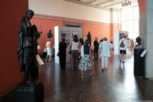 Wnętrze galerii sztuki z grupą ludzi zwiedzających wystawę. Na środku sali ustawione są duże białe ściany ekspozycyjne, na których wiszą w ramach kolorowe, nowoczesne fotografie lub plakaty. Między tymi ścianami przechadza się kilka osób w różnym wieku, większość to kobiety w letnich strojach. Po lewej stronie stoi na ciemnym postumencie rzeźba. Podłoga jest wyłożona płytkami z geometrycznym wzorem w odcieniach szarości z czerwonymi rombami. Ściany w tle mają ciepły, pomarańczowy kolor. Z prawej strony widać wysokie okna wpuszczające jasne, dzienne światło, które oświetla całą przestrzeń wystawową.