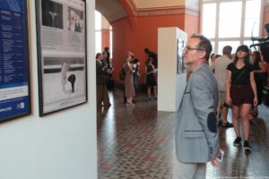 Wnętrze galerii sztuki z kilkoma osobami oglądającymi wystawę fotograficzną. Na pierwszym planie, po prawej stronie, stoi mężczyzna w szarej marynarce z ciemnymi łatami na łokciach i jasnych spodniach, trzyma w dłoni kilka kartek lub ulotek i zwraca się twarzą w stronę dużej, oprawionej fotografii wiszącej na ścianie po lewej. Na tej ścianie widać dwie duże, pionowe ramy: jedna z niebieskim panelem z tekstem, druga z kilkoma czarno-białymi zdjęciami i podpisami, w dolnej części tej fotografii przedstawiona jest osoba w płaszczu stojąca w śniegu obok psa. Podłoga ma geometryczny wzór z małych płytek w odcieniach szarości z czerwonymi rombami. W głębi sali, przy kolejnych białych panelach wystawowych, stoi kilka osób – część trzyma katalogi lub notatki, jedna kobieta w długiej, beżowej sukience ma na plecach plecak. Po prawej stronie, bliżej okna, idzie kobieta w czarnym T‑shirc ie i bordowych szortach, z małą torbą na pasku przez ramię. W tle widoczne są duże, wysokie okna, przez które wpada jasne światło, oraz ciemne rzeźby, w tym figura łucznika z napiętym łukiem. Ściany pomieszczenia są w ciepłym, ceglastym kolorze z jaśniejszym gzymsem pod sufitem.