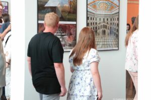 Para osób stojących przed ścianą z dużymi fotografiami. Na pierwszym planie stoi mężczyzna o jasnych, krótkich włosach, jasnej karnacji, w czarnej koszulce i jasnych spodniach, odwrócony tyłem. Obok niego, również tyłem, stoi kobieta o długich, rudawobrązowych włosach i jasnej karnacji; ma na sobie białą sukienkę w fioletowe kwiaty, przewiązaną w pasie, w ręce trzyma cienki, prostokątny przedmiot przypominający telefon. Przed nimi na ścianie wiszą duże, pionowe ramy ze zdjęciami; po lewej widać kilka zdjęć ułożonych w jednym kadrze, a po prawej jedno duże zdjęcie wnętrza budynku z arkadami i przeszklonym, ozdobnym sufitem w pomarańczowo-biały wzór. Po bokach kadru znajdują się fragmenty innych osób w jasnych ubraniach, sugerujące, że jest to zatłoczone wnętrze wystawy lub galerii.