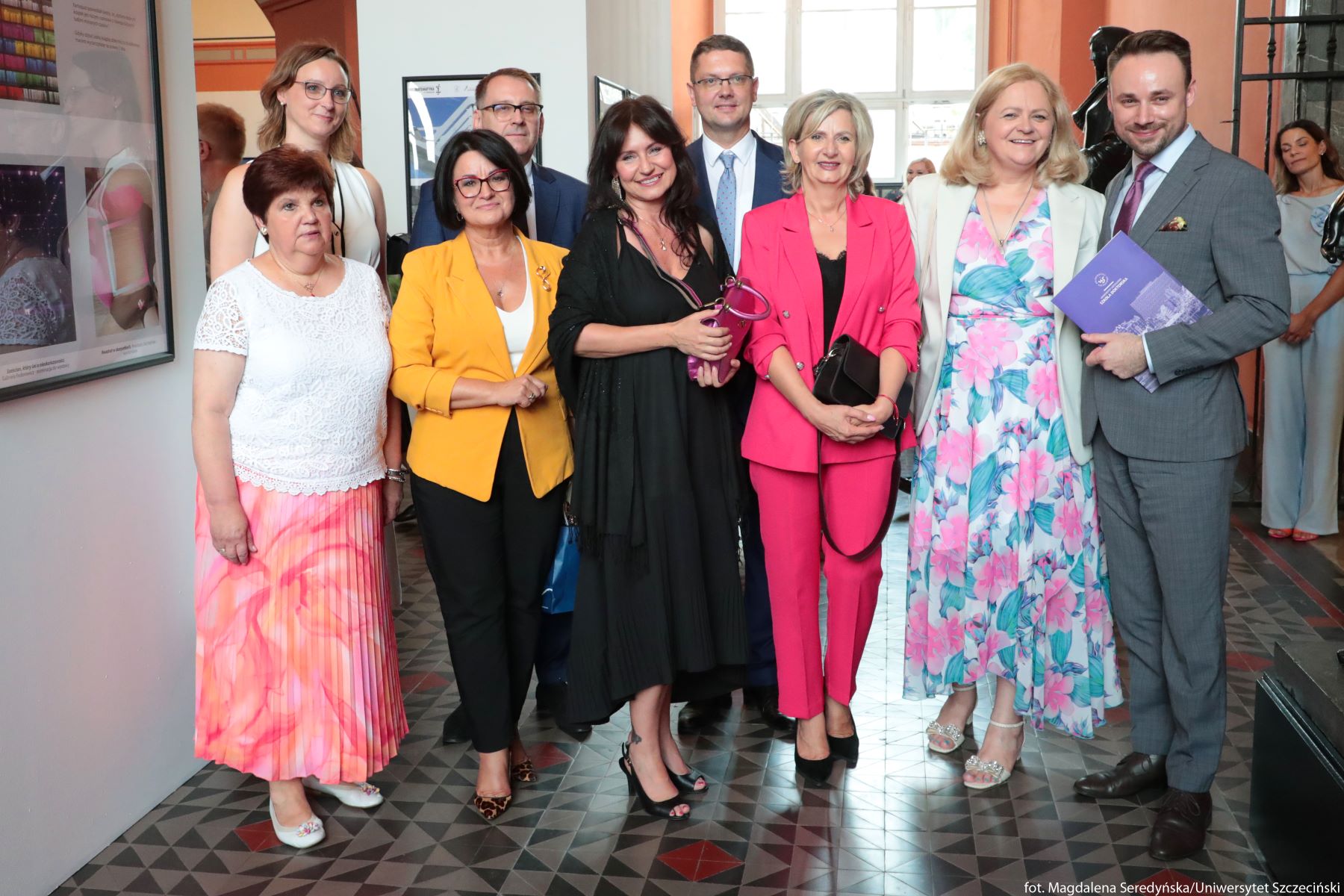 Grupa dziewięciu osób stojących razem we wnętrzu budynku na ozdobnej, kafelkowej podłodze. Wszyscy są dorośli, o jasnej karnacji, elegancko ubrani jak na uroczystość lub galę. Stoją w dwóch rzędach, zwróceni przodem do aparatu, większość uśmiechnięta. Z lewej strony znajduje się kobieta w białej koronkowej bluzce i długiej plisowanej spódnicy w odcieniach różu i pomarańczu, w białych butach na niewysokim obcasie. Obok niej stoi kobieta w żółtej marynarce, białym topie i czarnych, wąskich spodniach, w panterkowych czółenkach; ma krótkie, ciemne włosy i ciemne okulary w grubych oprawkach. Na środku, trochę z przodu, stoi kobieta w długiej czarnej sukience z dekoltem, na czarnych, wysokich sandałkach; w dłoniach trzyma różowo‑fioletową torebkę lub futerał na okulary. Po jej prawej stronie jest kobieta w wyrazistym, intensywnie różowym garniturze – marynarka i spodnie – z czarnym topem i czarną torebką na pasku, w czarnych czółenkach. Obok niej stoi kobieta w długiej, zwiewnej sukience w duży kwiatowy wzór w kolorach różu, błękitu i turkusu oraz w jasnej marynarce; ma jasne, półdługie włosy i jasne, ozdobne sandały na obcasie. Na skraju prawej strony stoi mężczyzna w jasnoszarym garniturze z białą koszulą i fioletowym krawatem; w dłoniach trzyma fioletową teczkę lub katalog. W drugim rzędzie za nimi widoczna jest kobieta w jasnej, bezrękawnikowej bluzce i długim naszyjniku, z jasnobrązowymi, do ramion włosami i okularami; obok niej stoi mężczyzna w okularach, granatowym garniturze i jasnej koszuli. Między osobami z pierwszego rzędu prześwituje jeszcze sylwetka mężczyzny w garniturze i jasnym krawacie. Tło stanowią jasne ściany z powieszonymi ramkami z plakatami lub zdjęciami; po lewej stronie wisi większa rama z kilkoma kolorowymi fotografiami. Z tyłu jest jasne okno wpuszczające dzienne światło oraz fragment rzeźby po prawej. W dalszym planie, po prawej stronie, stoi jeszcze jedna kobieta w jasnym stroju, częściowo zasłonięta. Na dole w prawym rogu widoczny jest biały napis z przypisem fotograficznym.