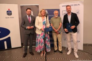 Od lewej: mężczyzna w szarym garniturze, kobieta w białej marynarce i długiej sukience w niebiesko-różowe kwiaty, która przemawia zza mikrofonu, mężczyzna w koszulce koloru khaki i kremowych spodniach, dalej wysoki mężczyzna w czarnej marynarce, błękitnej koszuli i białych spodnia. Za nimi rollupy PKO Banku Polskiego, Muzeum Narodowego w Szczecinie i Uniwizji – Telewizji Uniwersytetu Szczecińskiego.