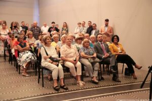 Grupa ludzi siedzących na krzesłach w pomieszczeniu. Na pierwszym planie kilka rzędów czarnych krzeseł, na których siedzą głównie starsze kobiety oraz dwóch elegancko ubranych dorosłych mężczyzn i jedna kobieta w żółtej marynarce. Osoby mają jasne i średnie karnacje, różne kolory włosów – od siwych po ciemne – i są ubrane raczej odświętnie, ale wygodnie: sukienki, bluzki, koszule, jasne spodnie, sandały i mokasyny. W środkowym rzędzie, po prawej stronie, siedzi mężczyzna w szarym garniturze z krawatem, obok niego kobieta w żółtej marynarce, białym topie i czarnych spodniach, w czarnych szpilkach. Przed nimi i obok nich siedzą starsze panie w jasnych bluzkach, swetrach i spodniach, niektóre z torebkami na kolanach. W tle, za siedzącymi, stoi zwarta grupa kilkudziesięciu osób – kobiety i mężczyźni w różnym wieku. Kilka osób ma założone ręce, ktoś trzyma aparat fotograficzny, jedna osoba patrzy w telefon. Ubrania są codzienne: koszulki, koszule, sukienki letnie, część osób ma okulary. Pomieszczenie ma gładkie, jasne ściany i jasne oświetlenie. Podłoga jest z ciemnych płytek z jaśniejszym wzorem w kratkę. Krzesła ustawione są w równych rzędach, tworząc wrażenie formalnego spotkania, wykładu lub uroczystości.