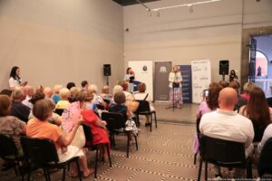 Sala wykładowa. Na pierwszym planie widownia tyłem do kadru. W tle kobieta w białej marynarce i długiej sukience w niebiesko-różowe kwiaty, która przemawia zza czarnego mikrofonu na statywie.