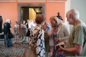Grupa osób w przestronnym wnętrzu o pomarańczowych ścianach i ozdobnej, geometrycznej posadzce. Scena wygląda jak spotkanie towarzyskie lub wernisaż – kilka osób stoi w małych grupach i rozmawia. Na pierwszym planie po prawej stronie stoi starszy mężczyzna o siwych włosach i brodzie, w jasnej, oliwkowej koszulce z krótkim rękawem; w dłoniach trzyma otwartą książkę lub broszurę. Obok niego, bardziej w środku, stoi kobieta o blond włosach do ramion, w jasnej marynarce i kolorowej, kwiatowej sukience; pochyla się w stronę niższej kobiety w oliwkowo‑zielonej bluzce, uśmiecha się szeroko, sprawia wrażenie zaangażowanej w rozmowę. Do tej małej grupy należy też kobieta plecami do obiektywu, w granatowej bluzce w duże jasne kwiaty, z beżową torebką przewieszoną przez ramię. W tle widać kilka innych osób, część ubrana elegancko, część bardziej swobodnie; stoją w korytarzu prowadzącym do jaśniej oświetlonego przejścia z otwartymi, metalowymi kratowanymi drzwiami. Po lewej i prawej stronie znajdują się ciemne rzeźby postaci ludzkich na cokołach, sugerujące, że miejsce może być muzeum lub galeria. Na dole po prawej widoczny jest mały biały napis z informacją o autorce zdjęcia i uczelni.