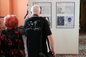 Dwoje dorosłych ludzi stoi przed białą ścianą z pracami wiszącymi w ramkach. Po lewej jest kobieta o krótkich, intensywnie rudych włosach, w sukience w ciemnym kolorze z dużym kwiatowym wzorem w czerwieniach i zieleniach, z czerwoną torebką na ramieniu. Po prawej jest mężczyzna z krótkimi siwymi włosami, w czarnej koszulce z białym nadrukiem na plecach; nadruk to ozdobny napis „Shoot To Frame” z kilkoma małymi okrągłymi symbolami nad nim. Na ramieniu ma przewieszoną czarną torbę na długim pasku. Przed nimi widać dwie duże pionowe ramy z czterema polami każda; w ramkach są fotografie i tekst, ale treści nie da się odczytać z powodu zbyt małej wielkości. W górnej części każdej ramy widnieją logotypy i napisy, a w dolnych częściach zdjęcia, między innymi jasnoszare z rozmytym kształtem, układ pól z cyframi oraz fotografia fioletowego, pióropuszowego kształtu przypominającego tancerkę w pozie z góry. Podłoga ma wzór w szare i białe geometryczne kafelki z czerwonymi akcentami. W tle po lewej stronie znajduje się fragment pomarańczowej ściany i ciemny fotel, a pośrodku, częściowo zasłonięta, ciemna rzeźba postaci.