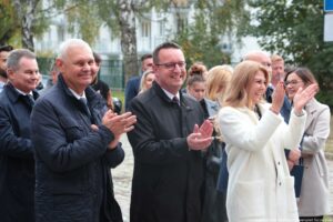 Otwarcie budynku Wydziału Prawa i Administracji Uniwersytetu Szczecińskiego. Na pierwszym planie trzy klaszczące osoby – dwóch mężczyzn i kobieta. W tle inni uczestnicy wydarzenia i drzewa.