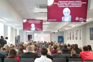 Otwarcie budynku Wydziału Prawa i Administracji Uniwersytetu Szczecińskiego. Ludzie siedzący w sali (tyłem do kadru). Pod sufitem dwa ekrany z białym napisem na bordowym tle: Uroczystość nadania imienia auli im. Profesora Bronisława Ziemianina Wydziału Prawa i Administracji Uniwersytetu Szczecińskiego. Poniżej czarno-białe zdjęcie profesora.