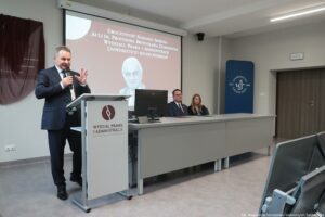 Otwarcie budynku Wydziału Prawa i Administracji Uniwersytetu Szczecińskiego. Ujęcie bokiem. Po lewej mównica, a z nią mężczyzna z mikrofonem, za nim na ścianie zasłonięta tablica pamiątkowa. W tle ekran z białym napisem na bordowym tle: Uroczystość nadania imienia auli im. Profesora Bronisława Ziemianina Wydziału Prawa i Administracji Uniwersytetu Szczecińskiego. Poniżej czarno-białe zdjęcie profesora.