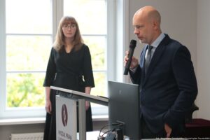 Mężczyzna w granatowym garniturze przemawiający do mikrofonu przy mównicy, obok laptopa i monitora komputerowego. Scena wygląda na salę wewnątrz budynku, z dużymi jasnymi oknami w tle. Po prawej stronie kadru stoi łysy mężczyzna o jasnej skórze, w białej koszuli, krawacie i ciemnym garniturze. Trzyma czarny mikrofon blisko ust i ma poważny wyraz twarzy; druga dłoń jest w kieszeni spodni. Przed nim stoi otwarty laptop oraz duży monitor ustawiony bokiem, z widocznymi kablami na dole. Po lewej stronie, w tle, stoi kobieta o jasnej skórze w czarnej sukience z długimi rękawami. Ma proste, jasnobrązowe lub ciemny blond włosy z grzywką i nosi okulary. Patrzy w kierunku mężczyzny lub przodu sali, z neutralnym wyrazem twarzy. Na froncie mównicy jest znak z grafiką i tekstem „WYDZIAŁ PRAWA”. W prawym dolnym rogu zdjęcia jest mały napis: „fot. Magdalena Seredyńska / Uniwersytet Szczeciński”.