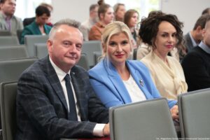 Grupa osób siedzących w rzędach na sali konferencyjnej lub wykładowej, z trzema osobami na pierwszym planie. Na pierwszym planie po lewej stronie siedzi mężczyzna o jasnej karnacji, z krótkimi siwymi włosami, w granatowej marynarce w kratę, białej koszuli i ciemnym krawacie. Ma lekki uśmiech i jest zwrócony twarzą w stronę aparatu. W środku siedzi kobieta o jasnej karnacji, z jasnymi blond włosami upiętymi z tyłu, w niebieskiej marynarce i białej bluzce. Ma czerwone usta i patrzy w stronę aparatu; na klapie marynarki jest przypięta mała ozdoba lub broszka. Po prawej siedzi kobieta o jasnej karnacji, z ciemnymi, kręconymi włosami upiętymi wysoko, w jasnej kremowej bluzce. Ma wyraz twarzy neutralny i również jest zwrócona w stronę aparatu. W tle widać kilka innych osób siedzących na szarych krzesłach, lekko nieostrych. W prawym dolnym rogu jest mały podpis fotograficzny: „fot. Magdalena Seredyńska/Uniwersytet Szczeciński”.