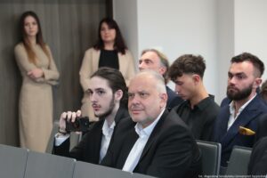 Otwarcie budynku Wydziału Prawa i Administracji Uniwersytetu Szczecińskiego. Ujęcie bokiem na ludzi siedzących w rzędach w sali. Na pierwszym planie młody mężczyzna nagrywający wystąpienie telefonem.