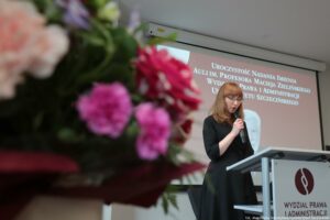 Uroczystość nadania auli im. Profesora Macieja Zielińskiego – Wydział Prawa i Administracji Uniwersytetu Szczecińskiego. Po lewej kwiaty. W tle po prawej mównica, a z nią kobieta z mikrofonem (ujęcie bokiem od dołu). W tle ekran z białym napisem na bordowym tle: Uroczystość nadania imienia auli im. Profesora Maciej Zielińskiego Wydziału Prawa i Administracji Uniwersytetu Szczecińskiego. Poniżej czarno-białe zdjęcie profesora.