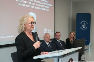Uroczystość nadania auli im. Profesora Macieja Zielińskiego – Wydział Prawa i Administracji Uniwersytetu Szczecińskiego. Po lewej mównica, a z nią kobieta z mikrofonem (ujęcie bokiem, do pasa). W tle ekran z białym napisem na bordowym tle: Uroczystość nadania imienia auli im. Profesora Maciej Zielińskiego Wydziału Prawa i Administracji Uniwersytetu Szczecińskiego. Poniżej czarno-białe zdjęcie profesora.