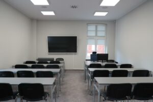 Otwarcie budynku Wydziału Prawa i Administracji Uniwersytetu Szczecińskiego. Sala wykładowa – krzesła, ławki, na ścianie po lewej czarny ekran, po prawej okno, a przed nim biurko z monitorem.