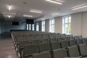 Otwarcie budynku Wydziału Prawa i Administracji Uniwersytetu Szczecińskiego. Sala wykładowa – rzędy ciemnoszarych składanych krzeseł. Dwa ekrany pod sufitem.