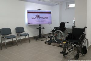 Otwarcie budynku Wydziału Prawa i Administracji Uniwersytetu Szczecińskiego. Sala symulacji. Na środku ekran na podstawie. Po prawej dwa wózki inwalidzkie. Po lewej dwa krzesła.