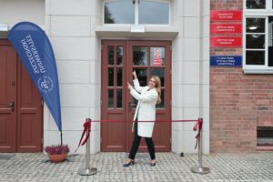 Otwarcie budynku Wydziału Prawa i Administracji Uniwersytetu Szczecińskiego. Wejście do budynku, przed którym jest czerwona wstęga. Za wstęgą dziekan Wydziału dr hab. Ewelina Cała-Wacinkiewicz, prof. US wskazująca palcami na drzwi. Po lewej granatowa flaga z białym logiem uczelni.