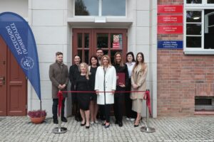 Otwarcie budynku Wydziału Prawa i Administracji Uniwersytetu Szczecińskiego. Wejście do budynku, przed którym jest czerwona wstęga. Za wstęgą pracownicy Wydziału i dziekan dr hab. Ewelina Cała-Wacinkiewicz, prof. US. Po lewej granatowa flaga z białym logiem uczelni.