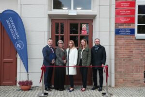 Otwarcie budynku Wydziału Prawa i Administracji Uniwersytetu Szczecińskiego. Wejście do budynku, przed którym jest czerwona wstęga. Za wstęgą dziekan Wydziału dr hab. Ewelina Cała-Wacinkiewicz, prof. US i prodziekani. Po lewej granatowa flaga z białym logiem uczelni.