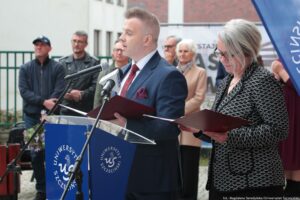 Otwarcie budynku Wydziału Prawa i Administracji Uniwersytetu Szczecińskiego. Ujęcie bokiem. Na pierwszym planie dwoje prodziekanów Wydziału stojących za granatową mównicą z białym okrągłym logiem US. W tle inni uczestnicy wydarzenia i drzewa.