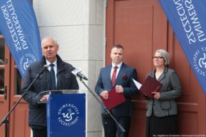 Otwarcie budynku Wydziału Prawa i Administracji Uniwersytetu Szczecińskiego. Na pierwszym planie za granatową mównicą z białym okrągłym logiem uczelni rektor US. Za nim po prawej dwoje prodziekanów Wydziału. W tle wejście do budynku.