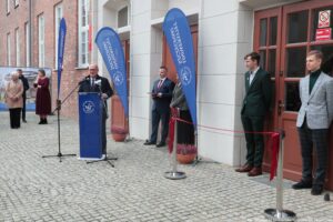 Otwarcie budynku Wydziału Prawa i Administracji Uniwersytetu Szczecińskiego. Po lewej za granatową mównicą z białym okrągłym logiem prezydent Szczecina. Obok dwoje prodziekanów Wydziału. Po prawej stronie czerwona wstęga na metalowych słupkach.