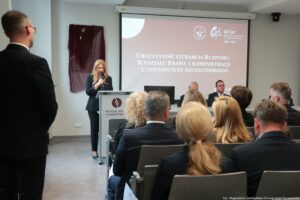 Ludzie siedzący w sali (tyłem do kadru). Na środku ekran z napisem uroczystość otwarcia budynku Wydziału Prawa i Administracji Uniwersytetu Szczecińskiego (w rogu logo uczelni i 40-lecia WPiA). Po lewej stronie dziekan WPiA dr hab. Ewelina Cała-Wacinkiewicz, prof. US za mównicą. Za nią zasłonięta tablica pamiątkowa.