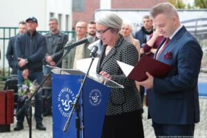 Otwarcie budynku Wydziału Prawa i Administracji Uniwersytetu Szczecińskiego. Ujęcie bokiem. Na pierwszym planie dwoje prodziekanów Wydziału stojących za granatową mównicą z białym okrągłym logiem US. W tle inni uczestnicy wydarzenia i drzewa.