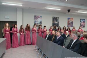 Otwarcie budynku Wydziału Prawa i Administracji Uniwersytetu Szczecińskiego. Ujęcie bokiem. Uczestnicy siedzący w rzędach w sali. W tle pod ścianą Chór Uniwersytetu Szczecińskiego – kobiety ubrane w ciemnoróżowe suknie.