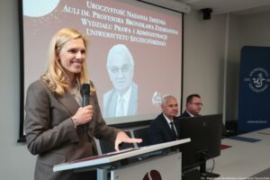 Otwarcie budynku Wydziału Prawa i Administracji Uniwersytetu Szczecińskiego. Po lewej mównica, a z nią kobieta z mikrofonem (ujęcie bokiem, do pasa). W tle ekran z białym napisem na bordowym tle: Uroczystość nadania imienia auli im. Profesora Bronisława Ziemianina Wydziału Prawa i Administracji Uniwersytetu Szczecińskiego. Poniżej czarno-białe zdjęcie profesora.