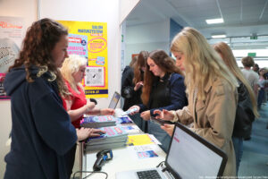 Po lewej bokiem dwie kobiety obsługujące stoisko, na którym leżą informatory i stoją laptopy z czytnikami. Po prawej osoby zainteresowane, skanujące kody QR. W tle żółty plakat z napisem Salon Maturzystów.