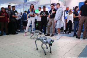 Na podłodze robot w kształcie psa. Za nim osoby sterujące robotem i trzymające informatory. W tle stoiska uczelni i uczniowie zainteresowani ofertą edukacyjną.