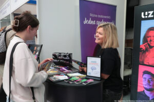 Po prawej bokiem kobieta obsługująca stoisko, na którym leżą informatory i ulotki. Po lewej zainteresowana uczennica trzymająca informator. W tle fioletowy rollup z napisem: Różne osobowości, jedna społeczność.