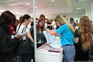 Po prawej bokiem kobieta obsługująca stoisko, na którym leżą informatory. Po lewej zainteresowane uczennice dopytujące o ofertę edukacyjną.