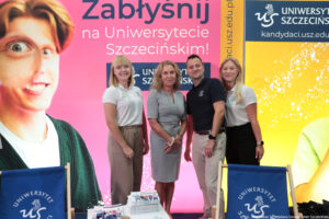 Troje pracowników Biura Rekrutacji (dwie kobiety i jeden mężczyzna) w polówkach z logiem Uniwersytetu Szczecińskiego stoją razem z prorektor ds. studenckich (druga od lewej) na tle różowej ścianki z napisem: Zabłyśnij na Uniwersytecie Szczecińskim. Przed nimi dwa leżaki z logiem US i informatory na stoliku.