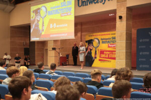 Ujęcie bokiem. Duża aula uniwersytecka. Na niebieskich fotelach siedzą uczniowie (tyłem). Na scenie prorektor ds. studenckich – dr hab. Katarzyna Kotarska, prof. US, która otwiera wydarzenie. Po lewej stronie od góry ekran z napisem: Szczeciński Salon Maturzystów – Perspektywy 2025, data wydarzenia 8–9 września i element graficzny – studentka z lupą, ubrana w togę i biret.